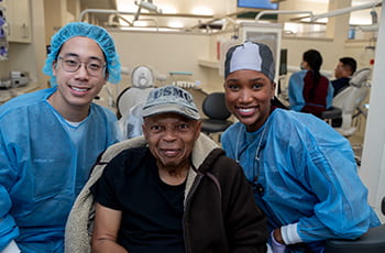 Two smiling dental students flank a veteran sitting in an operatory chair. 
